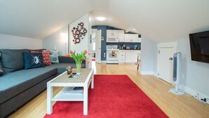 Een moderne woonkamer met een grijze bank, een witte salontafel en een rood kleed. In de achtergrond is een keuken met witte kasten en een magnetron zichtbaar.