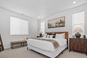 A bedroom with a large bed, a bench, a nightstand with a lamp, and a painting on the wall.
