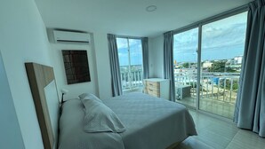 Ein modernes Hotelzimmer mit einem großen Bett, einem Nachttisch, einer Lampe und Blick auf die Stadt durch große Fenster.