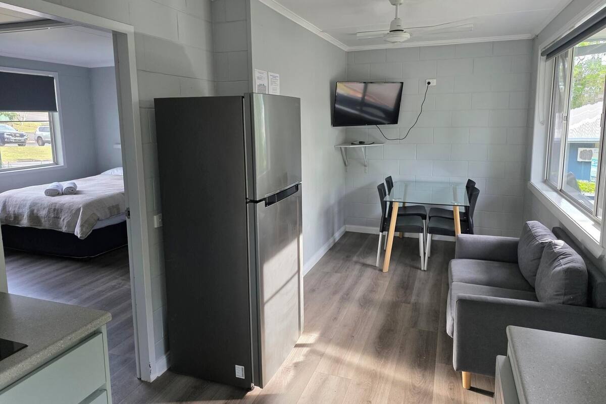 Una habitación compacta con cama, refrigerador, mesa de comedor con sillas, sofá y televisor montado en la pared.