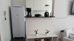 Cocina compacta con refrigerador, cafetera, licuadora y un pequeño estante blanco con adornos y frascos de almacenamiento.