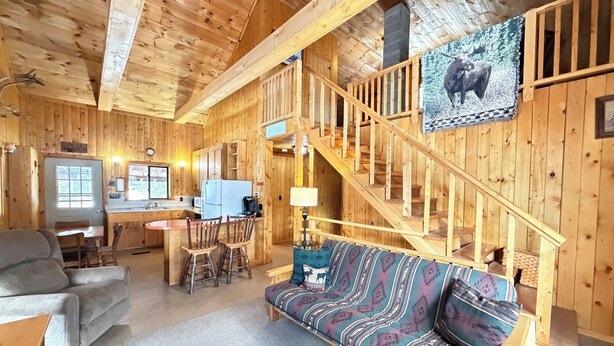 Interior d'una cabana de fusta amb un sofà, cadires, una taula de dinar, una zona de cuina i una escala.
