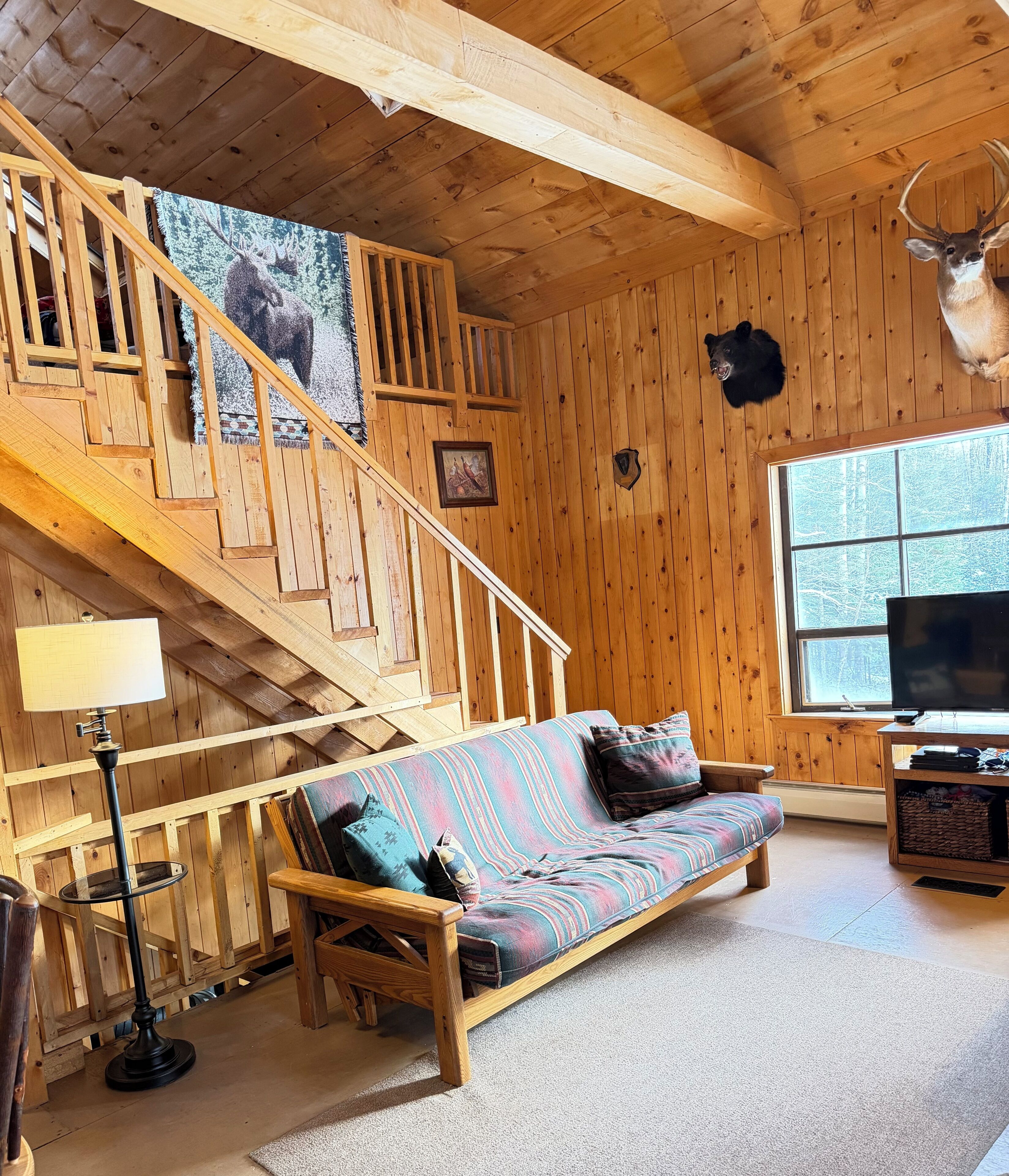 A wooden cabin interior with a sofa, a TV, a staircase, and a mounted deer head.