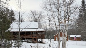 Una cabana de fusta amb un teulat cobert de neu, envoltada d'arbres i un petit cobert amb una bandera dels Estats Units.