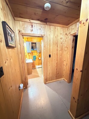 A wooden interior hallway leading to a kitchen area with a checkered window blind, a framed picture on the wall, and a visible light switch.