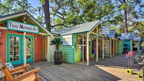 Une terrasse en bois avec une chaise en bois et un tonneau, entourée de bâtiments colorés portant les enseignes « Tipsy Mermaid Art » et « Island Gypsyseas ».