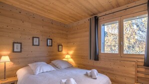 Een slaapkamer in een houten blokhut met een bed, twee bedlampjes, ingelijste foto’s aan de muur en een raam met uitzicht op bomen.