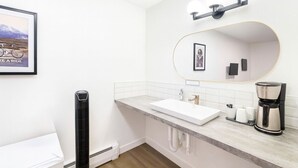 Une salle de bain moderne avec un lavabo blanc, un miroir avec éclairage intégré, un tableau encadré et une machine à café.