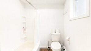 Une petite salle de bain blanche, équipée d’une douche, de toilettes et d’une étagère où sont rangés les articles de toilette.