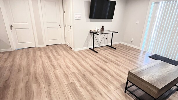 Een ruime kamer met houten vloer, een flatscreen televisie aan de muur, een glazen salontafel en een deur met een bordje.