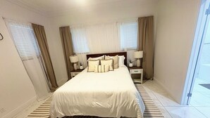 A hotel room with a large bed, two bedside tables with lamps, a window with curtains, and a striped rug. - BRAND NEW Austin Estates #3 2bed/2bath Apartment (Nassau)