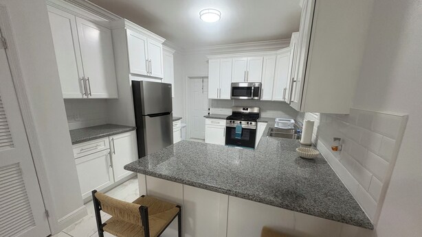A modern kitchen with white cabinets, a stainless steel refrigerator, a microwave, and a stove. There are two bar stools with woven seats. - BRAND NEW Austin Estates #3 2bed/2bath Apartment (Nassau)