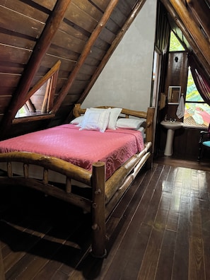 Ein Zimmer mit schrägen Wänden, in dem ein Holzbett, ein Fenster und ein Waschbecken zu sehen sind.