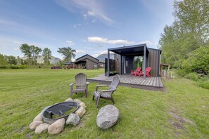 Eine moderne Außenterrasse mit zwei Adirondack-Stühlen, einer Feuerstelle und einem überdachten Sitzbereich mit roten Stühlen.