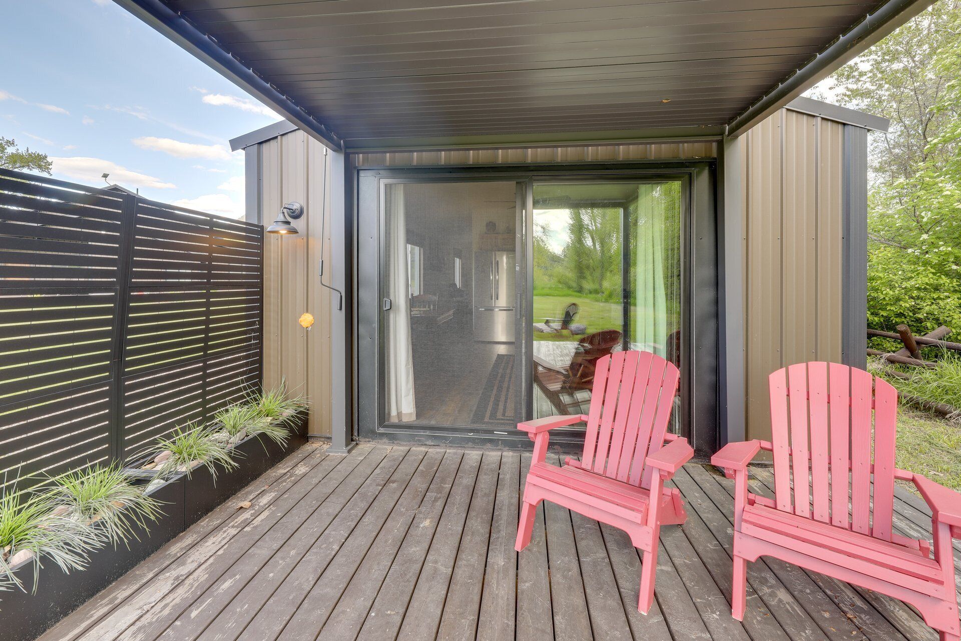 Een overdekt buitengebied met een schuifdeur naar een keuken, twee roze Adirondack stoelen en een houten terras.