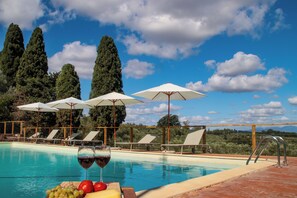 A poolside area with lounge chairs and umbrellas, a glass of red wine, and a fruit platter. - La Lunaria di Lajatico (Lajatico)