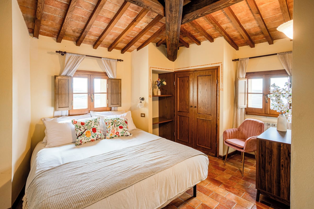 A bedroom with a large bed, wooden ceiling beams, a wooden wardrobe, and a chair.
