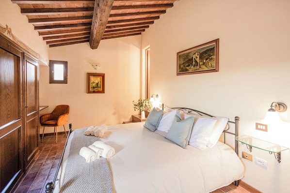 A bedroom with a bed, wooden ceiling beams, a chair, a nightstand with a lamp, and a painting on the wall. - La Lunaria di Lajatico (Lajatico)