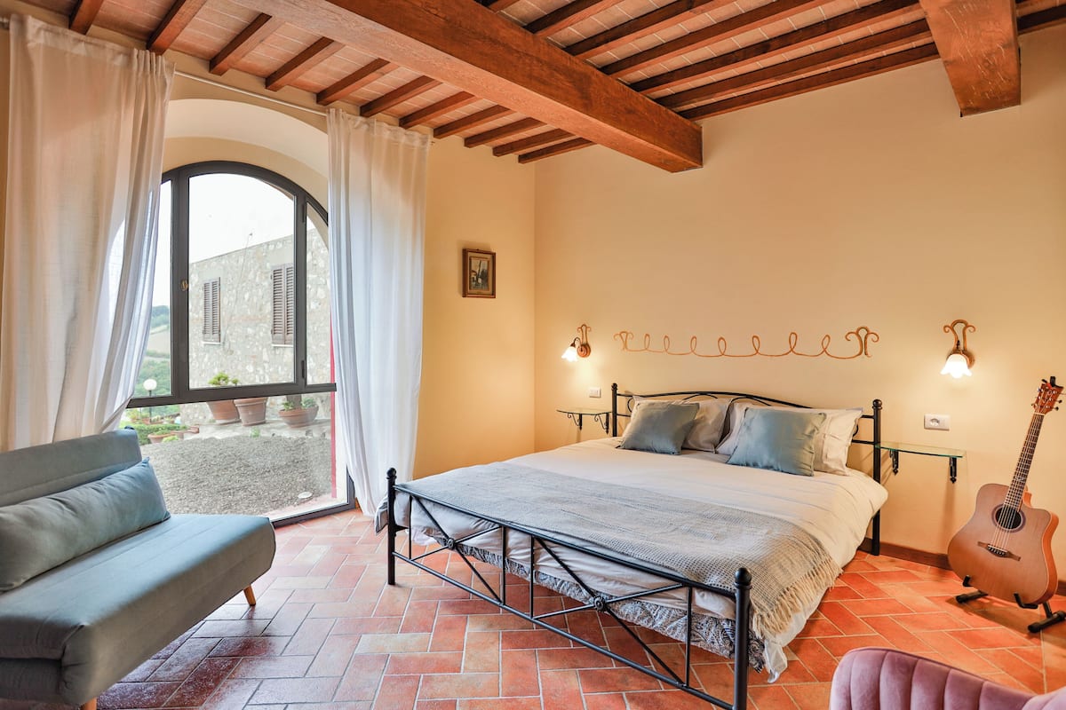 A bedroom with a bed, a guitar, a chair, and a view of the outside.