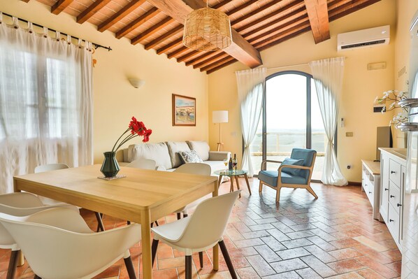 A spacious living area with a wooden dining table, white chairs, a sofa, and a blue armchair. There is a large window with a view, a hanging light fixture, and a white cabinet. - La Lunaria di Lajatico (Lajatico)