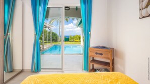 A bedroom with a bed, a wooden nightstand, and a view of a swimming pool and greenery through a glass door. - Coralyne (Les Trois-Ilets)
