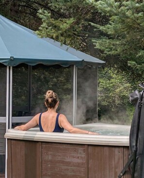 Una persona relajándose en un jacuzzi bajo una toldo, rodeada de árboles.