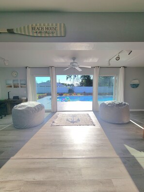 A spacious room with a large sliding glass door leading to a pool area, two bean bag chairs, a ceiling fan, and a rug with an anchor design. - Perfect 3-bedroom house with AC in charming Cape Coral (Cape Coral)