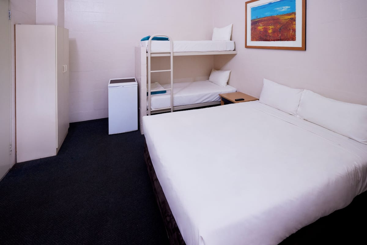 A hotel room with a bunk bed, a single bed, a small desk, a wardrobe, and a painting on the wall.
