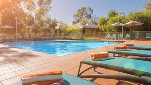A swimming pool with lounge chairs and umbrellas, surrounded by trees and a fence. - Outback Lodge (Ayers Rock)