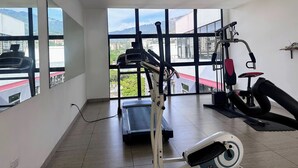 Un gimnasio con cinta de correr, máquina de pesas y un amplio ventanal que ofrece vistas a edificios y zonas verdes.