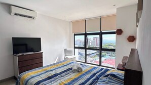 Un dormitorio moderno con una cama grande, un televisor en una cómoda, una ventana con vista a la ciudad y un ventilador de techo.