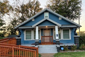 Una casa de dos pisos con fachada azul, un porche con barandilla de madera y un pequeño jardín delantero.