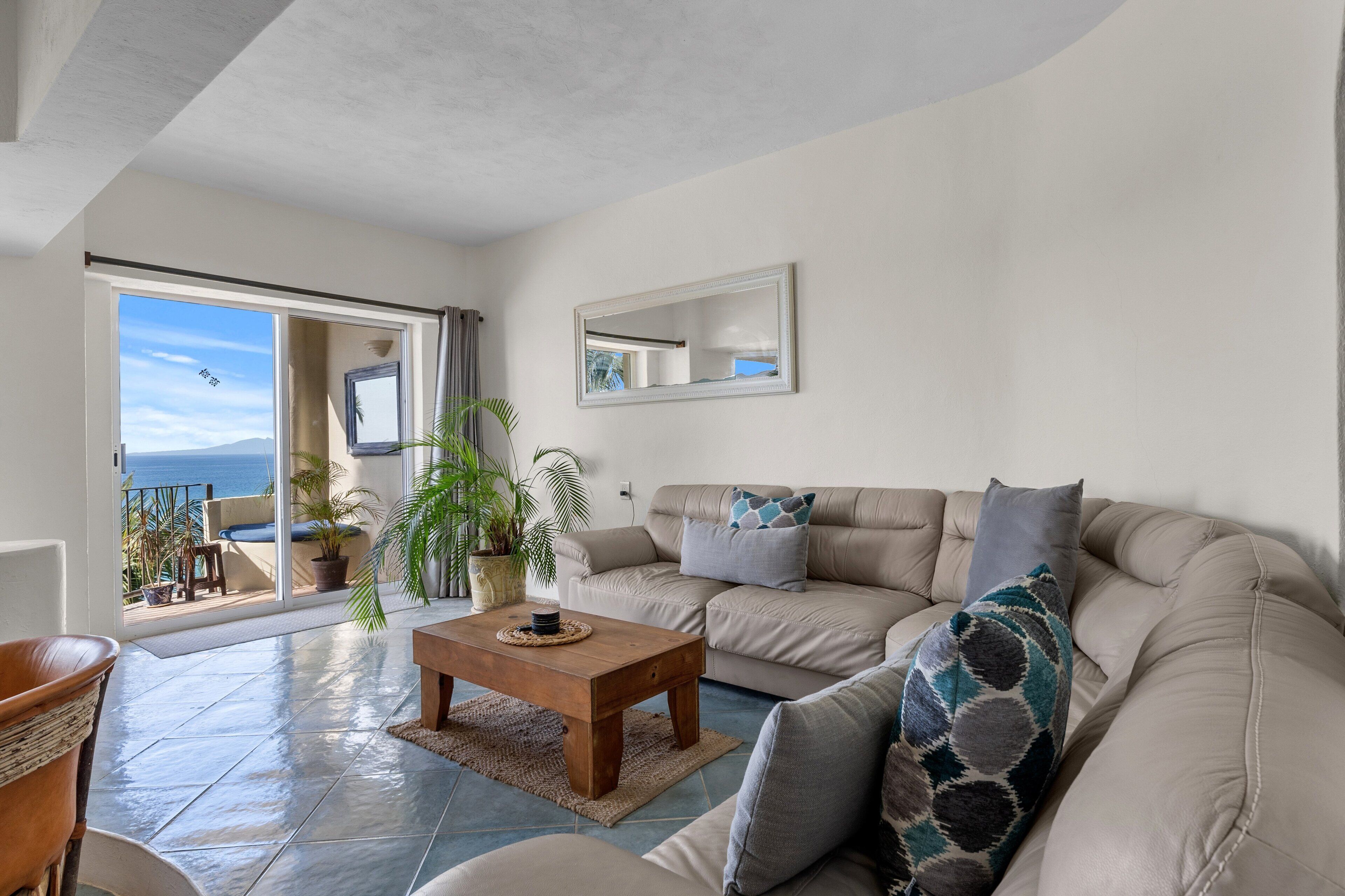 A living room with a beige sofa, a wooden coffee table, and a large window with a view of the sea.