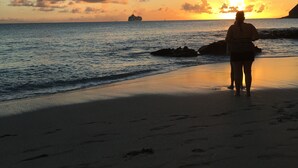 Una playa con una persona de espaldas al atardecer, un barco a lo lejos y formaciones rocosas.