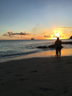 A beach with a person standing facing the sunset, a ship in the distance, and rocky outcrops. - Charming 1-bedroom studio in beautiful Sint Maarten. (Philipsburg)