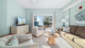Una sala de estar con un sofá color beige, un sillón blanco, una mesa de centro y un televisor sobre un mueble. Hay un balcón con vistas a palmeras y un edificio.