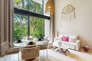 A modern living room with a dining area, large windows, and a white sofa with pink pillows. - Boho-Lux Loft in Mistiq Temple II. Rooftop Pool. 10 min from the beach. (Tulum)