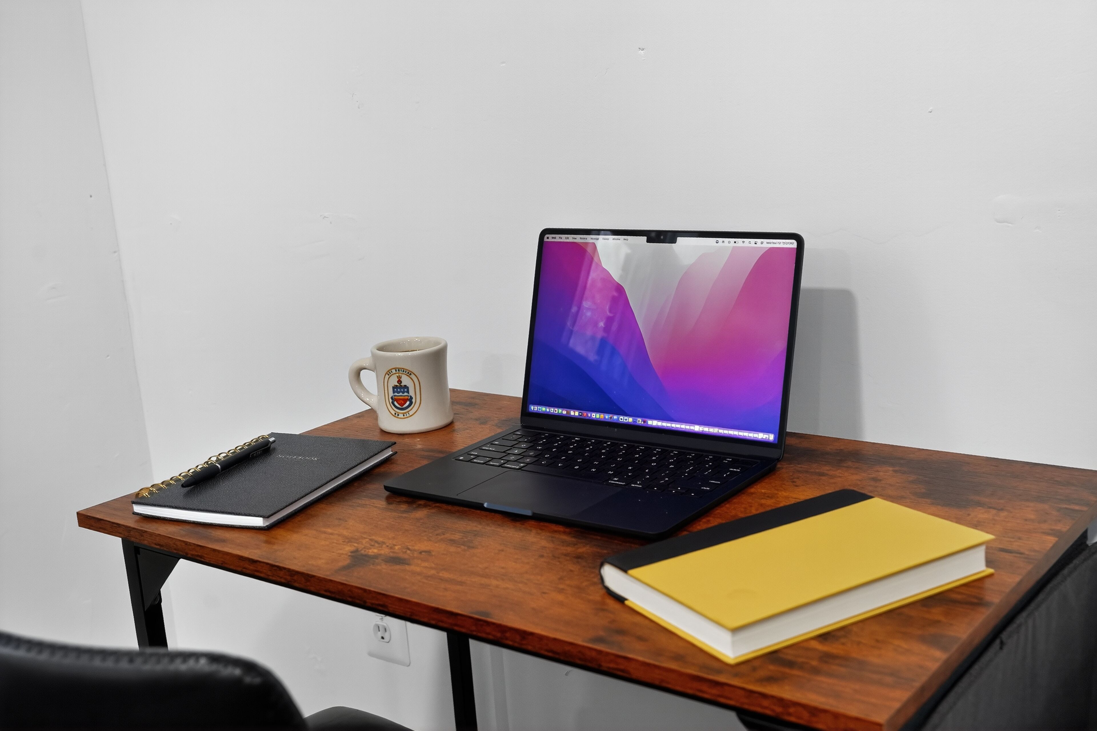 Un ordinateur portable sur un bureau en bois, avec un carnet, une tasse et un livre.