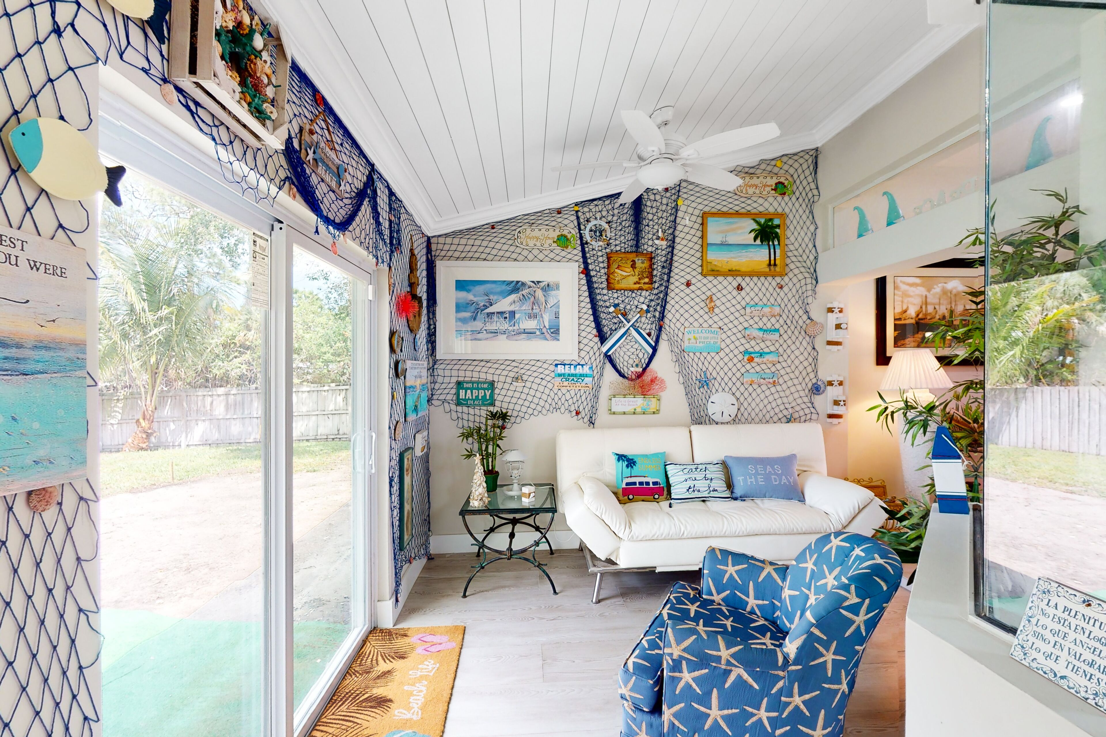 A room with a white sofa, a blue patterned chair, and a ceiling fan. There are framed pictures on the walls, a small table with a lamp, and a view of the outdoors through large windows.