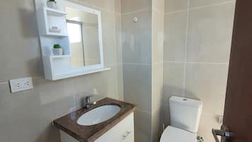 Un baño con tocador blanco, espejo, inodoro y repisa con plantas.