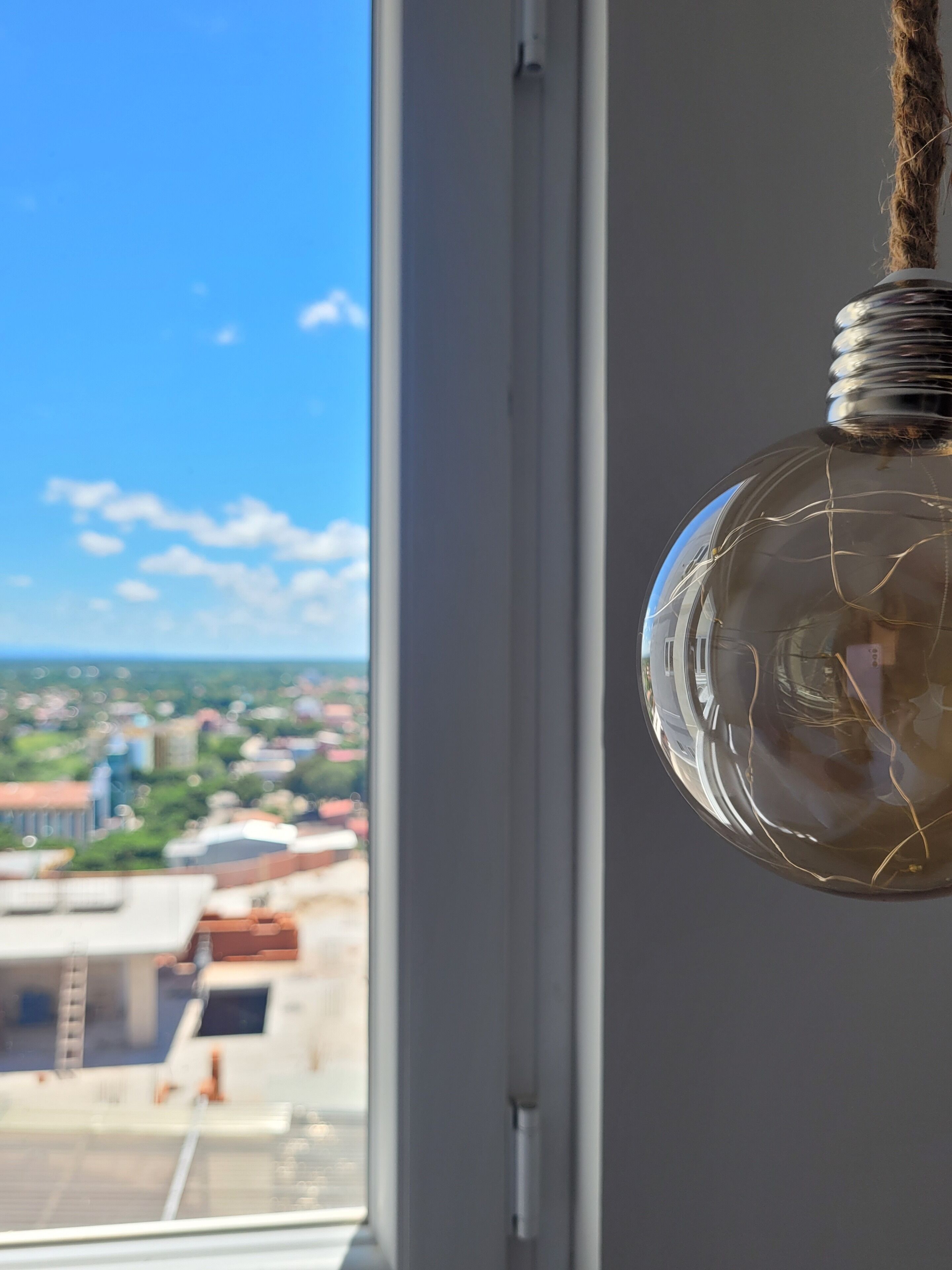 A hanging light bulb with visible internal filaments, viewed through a window overlooking a cityscape.