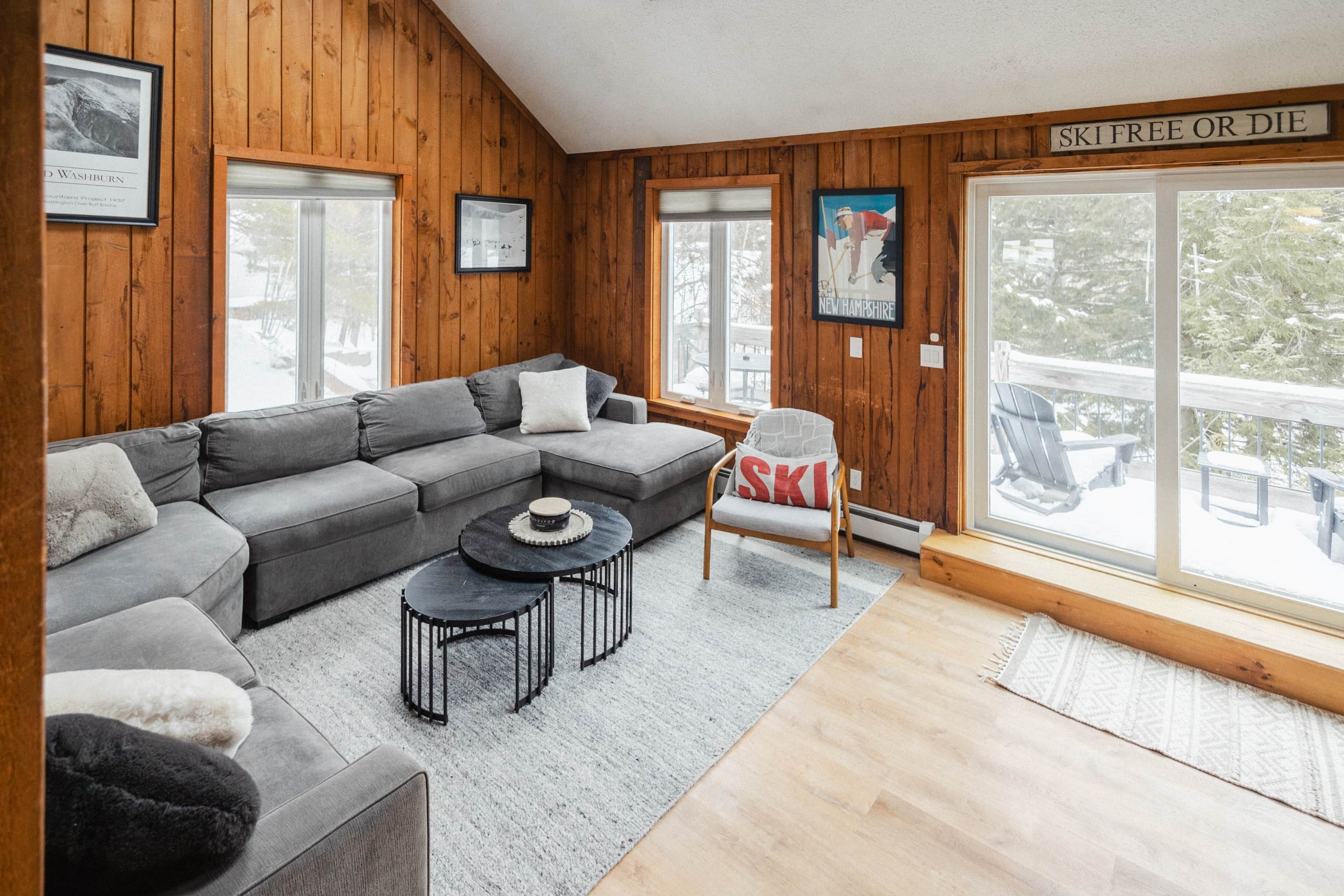 A cozy living room with a gray sectional sofa, a small round table, and a chair with a ski-themed pillow.