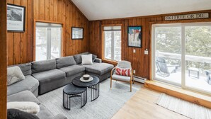 A cozy living room with a gray sectional sofa, a small round table, and a chair with a ski-themed pillow. - 1 Min to Cranmore - Sleeps 12 - 4 levels - Arcade Bunk Room- 3 Min to Downtown (North Conway)