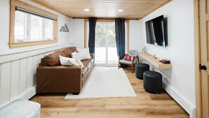 A cozy living room with a brown sofa, a white rug, a TV mounted on the wall, and a wooden ceiling. - 1 Min to Cranmore - Sleeps 12 - 4 levels - Arcade Bunk Room- 3 Min to Downtown (North Conway)