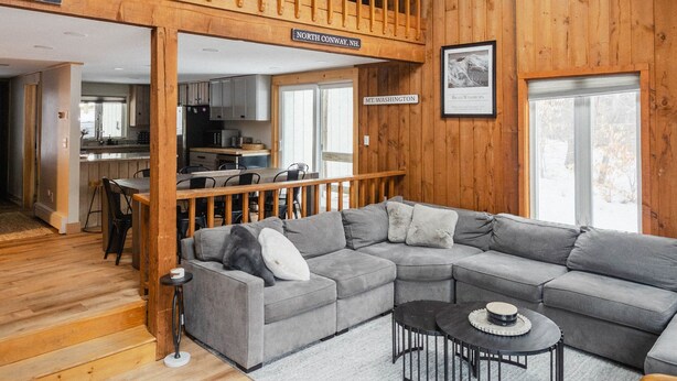 A spacious loft with a large gray sectional sofa, a dining area, a kitchen, and a wooden staircase. - 1 Min to Cranmore - Sleeps 12 - 4 levels - Arcade Bunk Room- 3 Min to Downtown (North Conway)