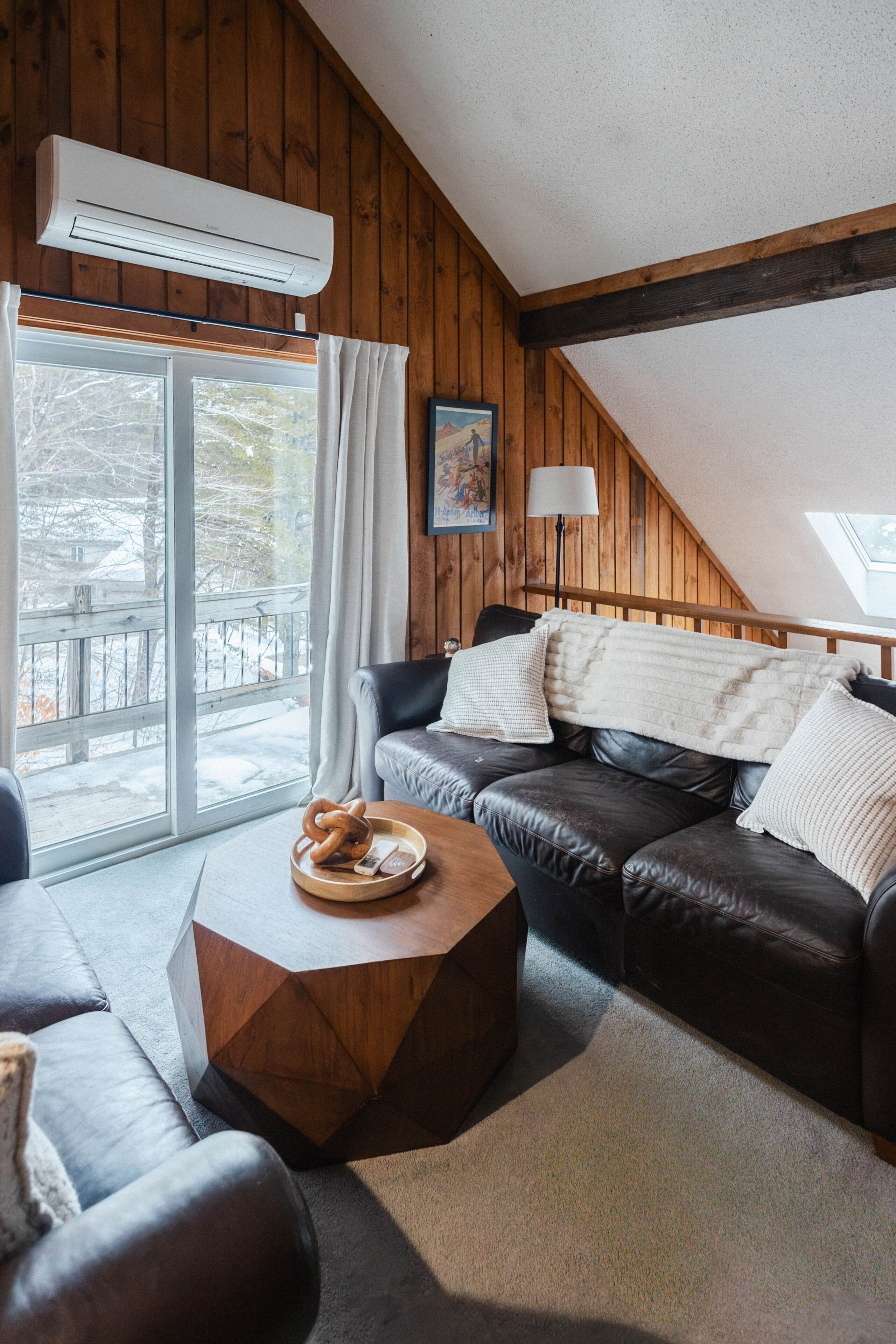 A cozy room with a leather sofa, a round wooden coffee table, and a large window with a view of a snowy deck.