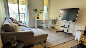 Ein modernes Wohnzimmer mit einer Couch, einem kleinen Tisch mit Pflanze, einem Fernseher auf einem Schrank und Blick ins Freie.