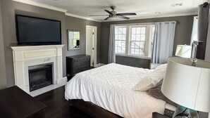 Quarto com cama, TV acima da lareira, ventoinha no teto e janela com cortinas.