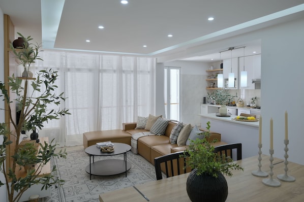 A modern living room with a sofa, coffee table, and dining table. There is a kitchen area with cabinets and a window.