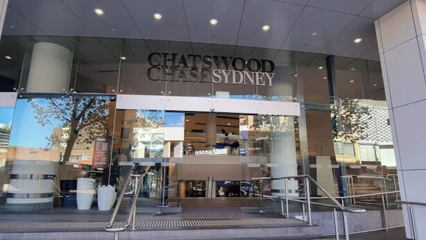 L'ingresso di Chatswood Chase Sydney, caratterizzato da una moderna facciata in vetro, posti a sedere visibili all'interno e una chiara vista sulla zona circostante.